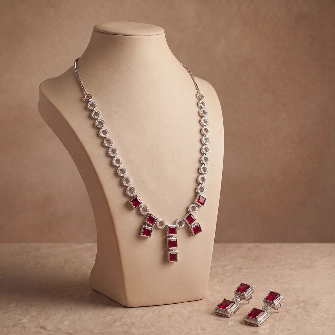 Silver-tone necklace and matching drop earrings with rectangular red gemstones, displayed on a beige jewelry bust against a warm brown studio background.