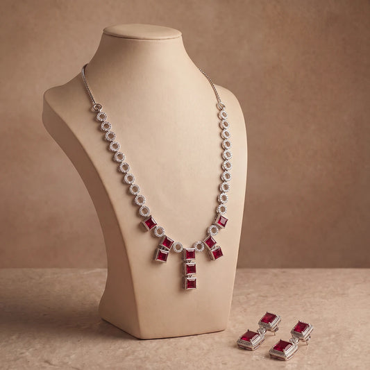 Silver-tone necklace and matching drop earrings with rectangular red gemstones, displayed on a beige jewelry bust against a warm brown studio background.