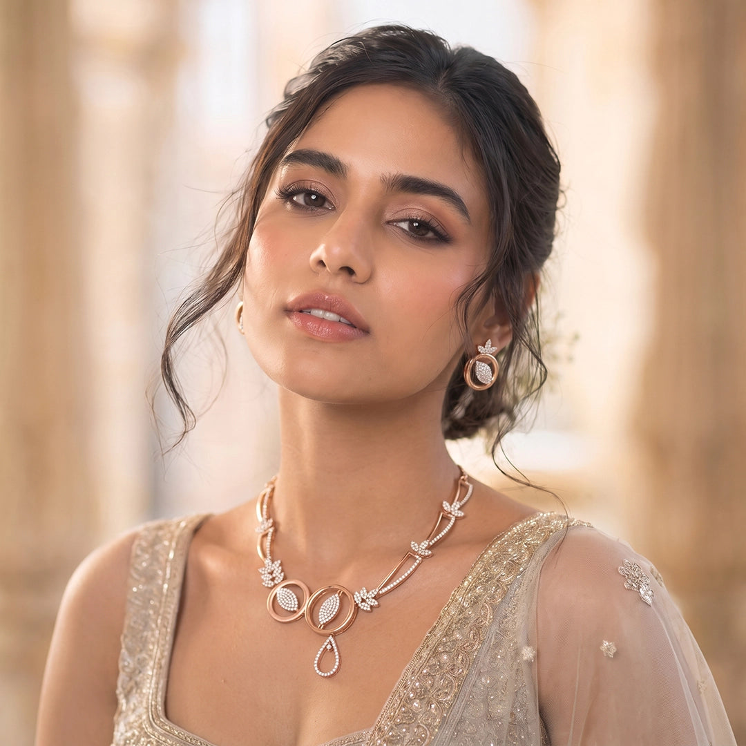 A woman wearing a delicate rose gold necklace with floral and teardrop motifs, paired with matching rose gold hoop earrings and a beige embellished traditional outfit, set against a softly blurred classical column background.
