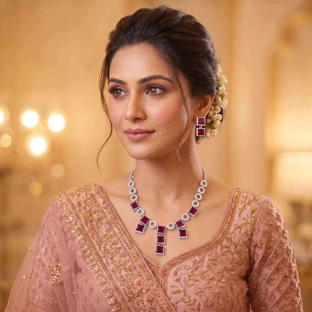A woman wearing a pink traditional outfit with gold embroidery, styled with a silver and ruby necklace and matching earrings, set against a warm, softly blurred background.