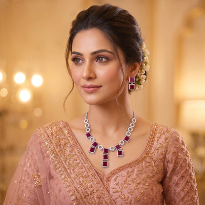 A woman wearing a pink traditional outfit with gold embroidery, styled with a silver and ruby necklace and matching earrings, set against a warm, softly blurred background.