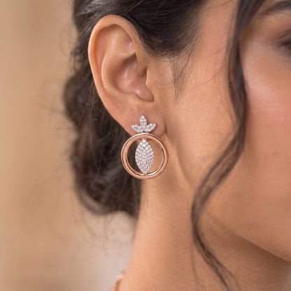 A close-up of a woman's ear wearing a rose gold hoop earring with a leaf-shaped stud set with clear crystals.