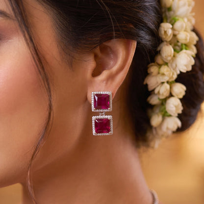 A close-up of a woman's ear wearing a pair of square drop earrings with deep pinkish-red gemstones in silver settings, adorned with small clear stones, with white flowers in her hair against a warm golden background.